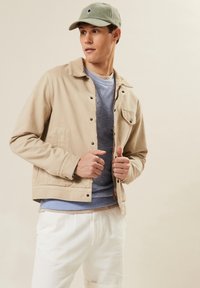 Beige Baumwolljacke mit Druckknöpfen, mit Taschendetails, über einem hellblauen Sweatshirt getragen, kombiniert mit weißen Hosen und einer grünen Mütze.