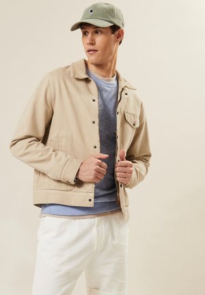 Beige katoenen jacket met drukknoopsluitingen, zakdetail, gelaagd over een lichtblauwe sweatshirt, gecombineerd met witte broek en een groene pet.