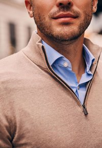 Beige ritsweater over een lichtblauwe overhemd met knopen. Heeft een gebreide textuur en een contrasterende rits. Focus op de halslijn en stofdetails.