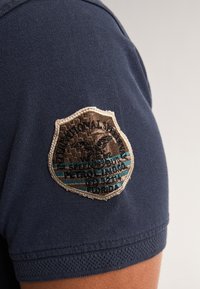 Námořnická modrá polo košile s texturovanou tkaninou a vyšitým nášivkem na rukávu s zlatým a tyrkysovým barevným schématem.