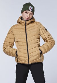 Tan puffer jacket with a hood, featuring black zipper and pocket accents. Ribbed green knitted beanie with a logo patch. Smooth, quilted texture.