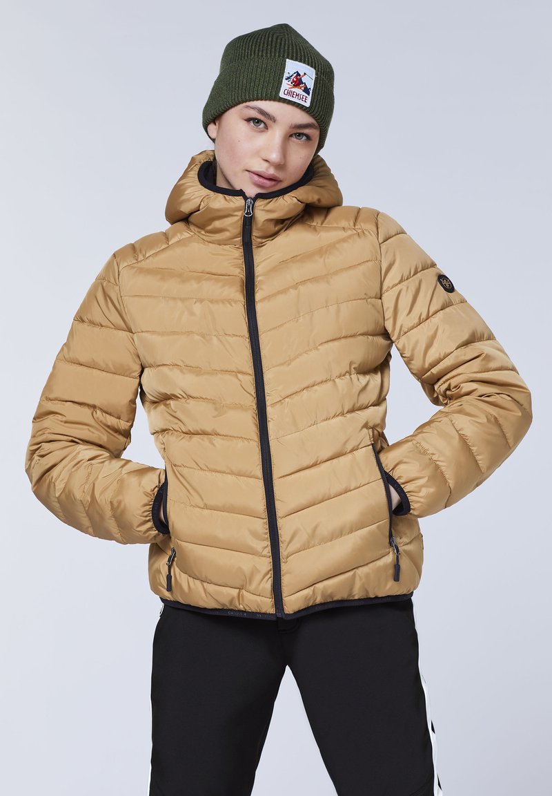 Tan puffer jacket with a hood, featuring black zipper and pocket accents. Ribbed green knitted beanie with a logo patch. Smooth, quilted texture.