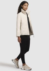 Chaqueta de plumón acolchada color crema con cuello alto, bolsillos laterales y cremallera frontal, combinada con leggings negros y zapatillas color beige.