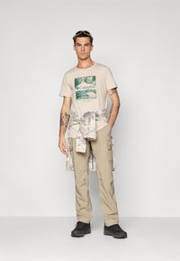 Beige t-shirt med grafik af et grønt bjerg, parret med lyse khaki bukser og sorte udendørssko. En lys tie-dye skjorte er bundet om taljen.