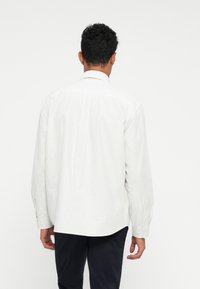 Uomo di spalle che indossa una camicia bianca a maniche lunghe con sottili righe verticali e pantaloni scuri, su uno sfondo chiaro e uniforme.