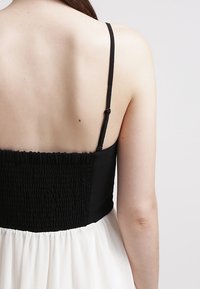 Zwart geplooid lijfje met dunne bandjes, overgaand in een vloeiende witte rok. Gladde textuur, zachte stof en minimale details.