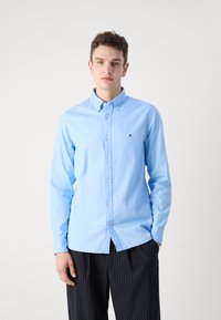 Camicia azzurro chiaro con colletto, slim fit e piccolo dettaglio del logo. Indossata con pantaloni a righe sottili. Texture liscia.