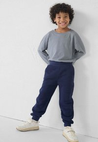 Grijze lange mouwen shirt met een ontspannen pasvorm, gecombineerd met marineblauwe trainingsbroek. Witte sneakers maken de outfit af. Effen achtergrond.