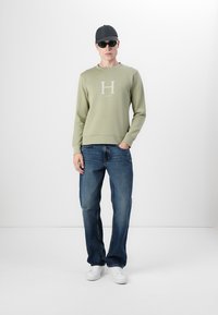 Sudadera verde clara con un logo blanco de "H", combinada con jeans oscuros y zapatillas blancas. El modelo lleva gafas de sol y una gorra negra.