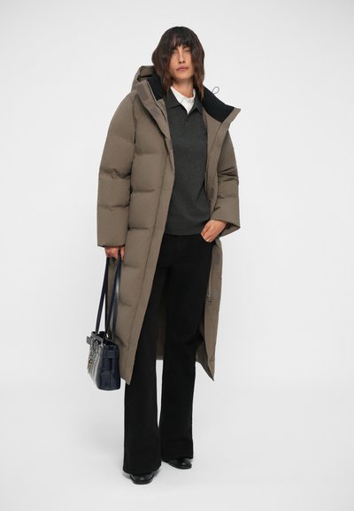 Manteau doudoune long, brun clair, avec capuche, présentant une fermeture éclair à l'avant et de grandes sections matelassées. Porté par-dessus un pull gris et un pantalon noir.