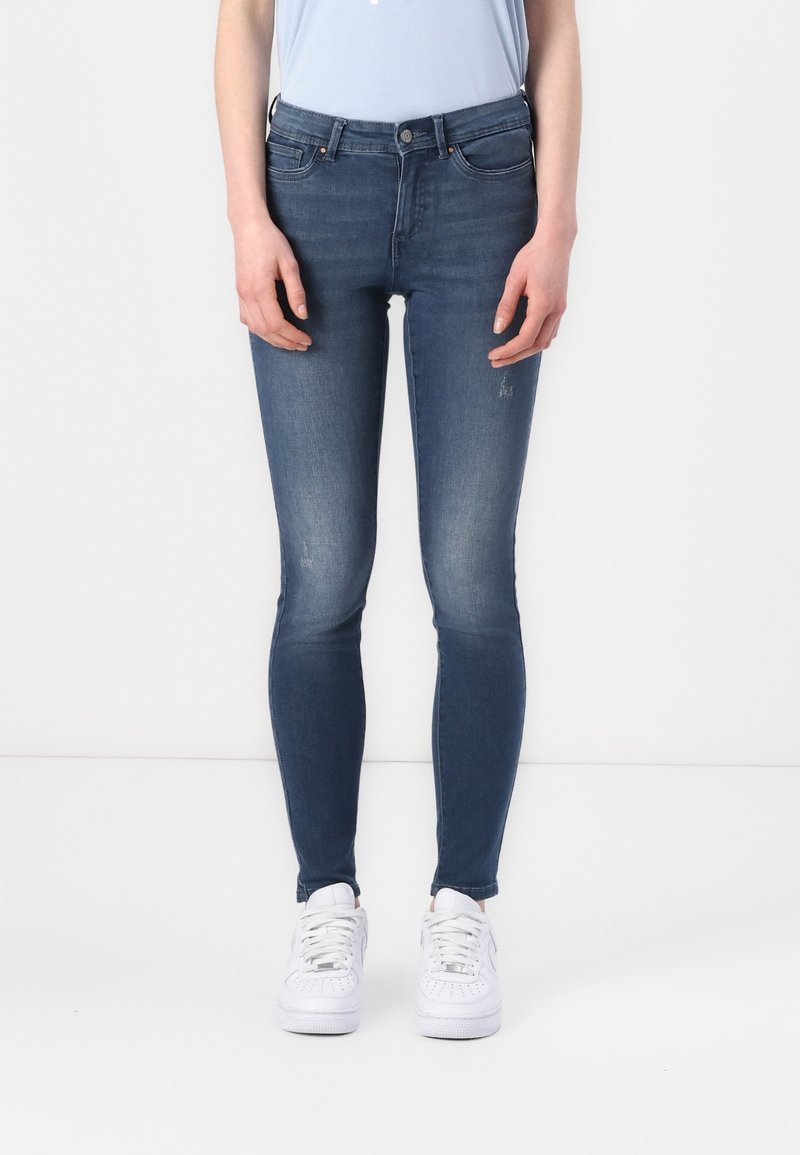 Personne portant un jean en denim bleu ajusté et des baskets blanches, debout devant un fond blanc uni.