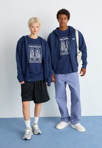 Sweatshirts da marinha sobre t-shirts gráficas, calções pretos e calças azul claro. Ténis em cinza e branco. Design simples e limpo com mínimos acentos.