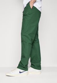 Green cotton pants with a relaxed fit, featuring a small Nike logo embroidered on the thigh and paired with white sneakers.