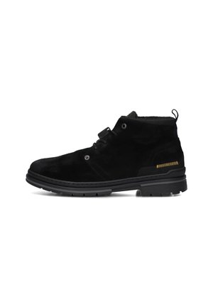 Bottes chelsea en suède noir avec semelle en caoutchouc, œillets latéraux et tirant. Présente un détail de surpiqûre jaune sur le talon pour le contraste.