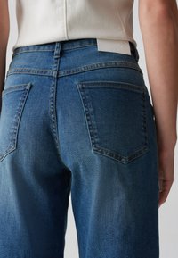 Blå denimjeans med en åtsittande midja, två bakfickor och en strukturerad yta. Taggen synlig vid bakre midjelinjen.
