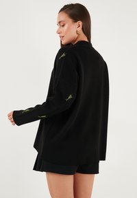 Crni oversized pulover s zelenim cvjetnim vezom na rukavima. Ima ležeran kroj i mekani materijal, dizajniran za opuštenu fit.