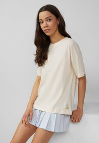 T-shirt en coton surdimensionné couleur crème avec des manches courtes, mettant en avant un petit patch graphique multicolore à l'ourlet, associé à une jupe plissée bleu clair.