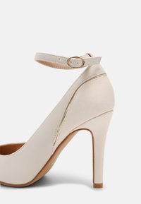 Chaussure à talon haut beige avec une finition texturée, une bride autour de la cheville et des accents en métal doré. Présente un bout pointu et un talon aiguille fin.