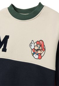 Felpa crema e blu navy con colletto a costine verde. Presenta una grafica colorata di Mario e una "M" nera sul davanti. Morbido materiale in cotone.