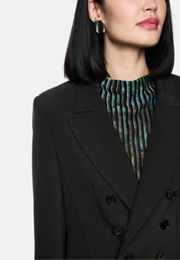 Schwarzes doppelt geknöpftes Blazer mit Revers, ausgestattet mit schwarzen Knöpfen. Darunter befindet sich ein mehrfarbig gestreiftes Oberteil mit hohem Kragen und unterschiedlichen Texturen.