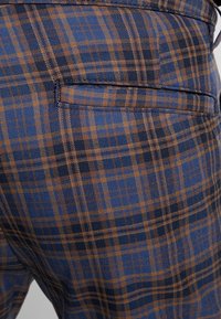 Blå och brun rutiga byxor med en strukturerad tygfinish och en bakficka. Rutmönstret har kvadrater i olika storlekar.