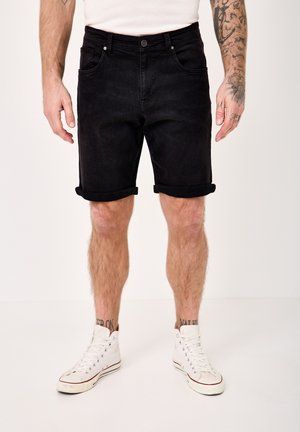 Man wearing black rolled-up denim shorts, white high-top sneakers, and a white shirt, standing with tattooed arms visible.