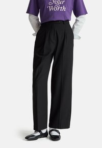 Zwarte geplooide broek met een recht model, gemaakt van zachte stof. Gecombineerd met zwarte schoenen en witte sokken.