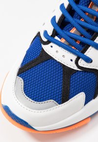 Scarpa da ginnastica atletica con tomaia in mesh blu, dettagli in pelle bianca, particolari neri e rifiniture arancioni. Presenta superfici testurizzate e lacci.