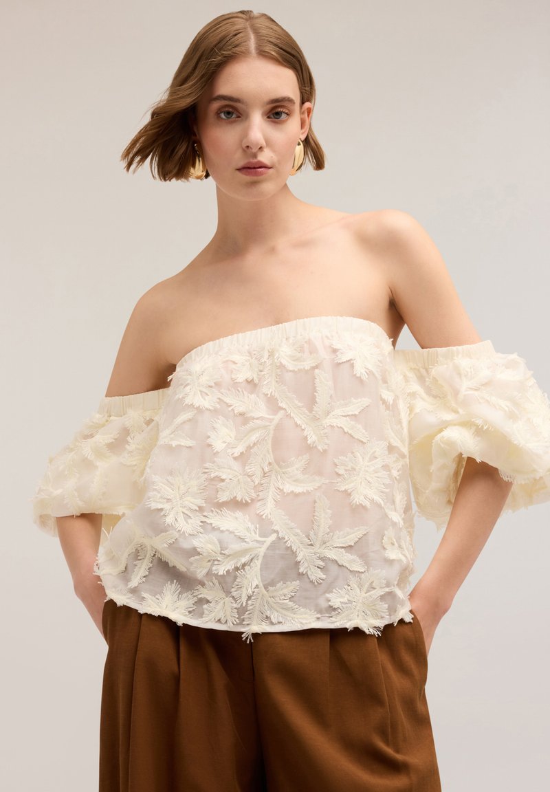 Blusa off-the-shoulder color crema con ricami floreali testurizzati, sovrapposizione trasparente e maniche corte a sbuffo, abbinata a pantaloni larghi marroni.