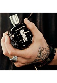 Flacon de parfum en forme de poing noir avec un capuchon métallique et le logo "DIESEL" en argent. Tenue dans une main ornée de tatouages et de bracelets.