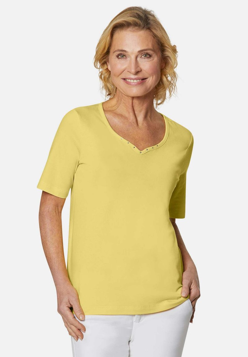 Gelbes Kurzarm-V-Ausschnitt-T-Shirt mit Verzierungen am Ausschnitt, aus weichem Stoff. Das Model trägt dazu weiße Hosen und lächelt.