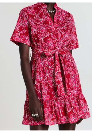 Frau trägt ein rotes Wickelkleid mit floralem Muster, kurzen Ärmeln und einem gebundenen Taillengürtel, accessoriert mit klobigen silbernen Ringen und einer Halskette.