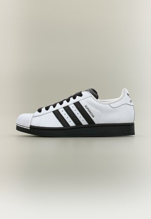 Baskets blanches Adidas Superstar avec des bandes noires, des lacets noirs et une semelle noire, montrées de profil sur un fond neutre.