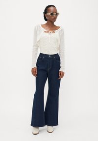 Lightweight white knit wrap top over a patterned off-white shirt, paired with high-waisted dark denim flared jeans and cream ankle boots.