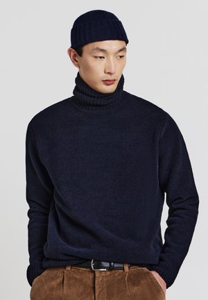Homme portant un pull col roulé bleu marine et un bonnet, avec un pantalon en velours côtelé marron et une ceinture noire, les mains dans les poches, regardant sur le côté.