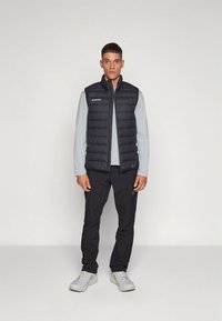 Črna puhasta telovnik z zadrgo spredaj, vodoravno prešivanje in logotipom. Nošen čez svetlo sivo majico z dolgimi rokavi ter kombiniran s črnimi hlačami in supergami.