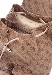 Bruine geblokte stoffen tas met een trekkoordsluiting, een binnenzak met rits en 'Guess' logo-accenten. Textuur oppervlakte met stikdetails.