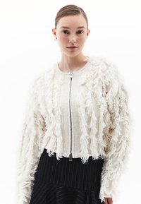 Cardigan corto color avorio con texture frangiata. Caratterizzato da una zip frontale e polsini a coste. Indossato sopra una gonna nera a righe.