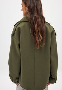 Olivza zaļa pārmēru mēteļi ar teksturētu apdari, ar platu apkakli, pogām pie piedurknēm un dekoratīvām plecu siksniņām.