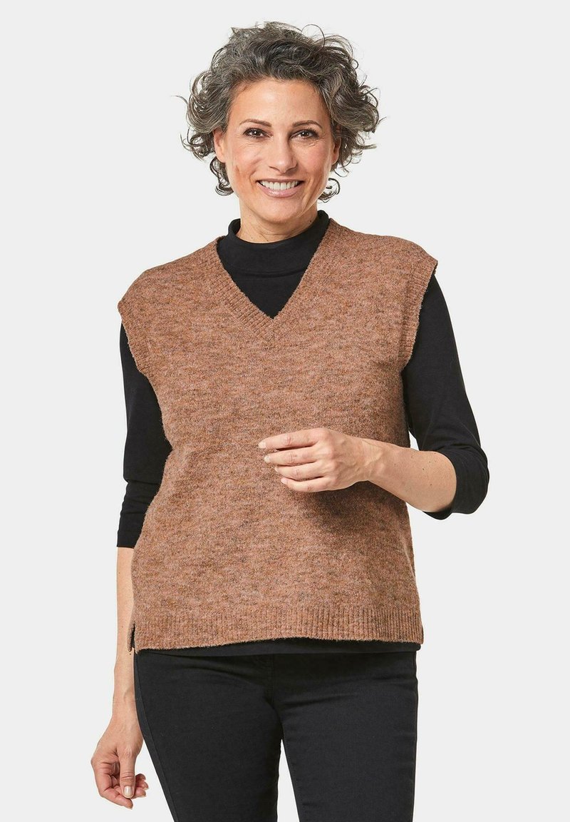 Bruine gebreide vest met een V-hals en korte mouwen, gedragen over een zwart langemouwen top. De stof heeft een zachte textuur en een subtiel patroon.