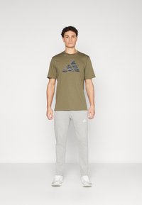Maslinasto zelena majica s crnim kamuflažnim Adidas logotipom, u kombinaciji svijetlosivim trenirkama s bijelim bočnim prugama i bijelim logotipom.