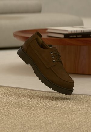 Καφέ καστόρινο κράνος με κορδόνια που αιωρείται πάνω από ένα μπεζ υφασμάτινο χαλί με ένα ξύλινο τραπεζάκι του καφέ και βιβλία στο παρασκήνιο.