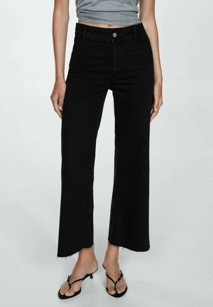 Frau trägt hoch taillierte schwarze Jeans mit weitem Bein und ausgefransten Säumen, kombiniert mit einem grauen Crop-Top und schwarzen, riemchen Sandaletten mit Absatz.