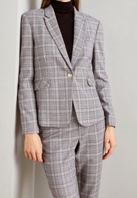 Szara kratkowana marynarka z złotym guzikiem, z kołnierzem z wycięciem i przednimi kieszeniami. Noszona na ciemnym golfie, w zestawie z pasującymi do niej kratkowanymi spodniami.