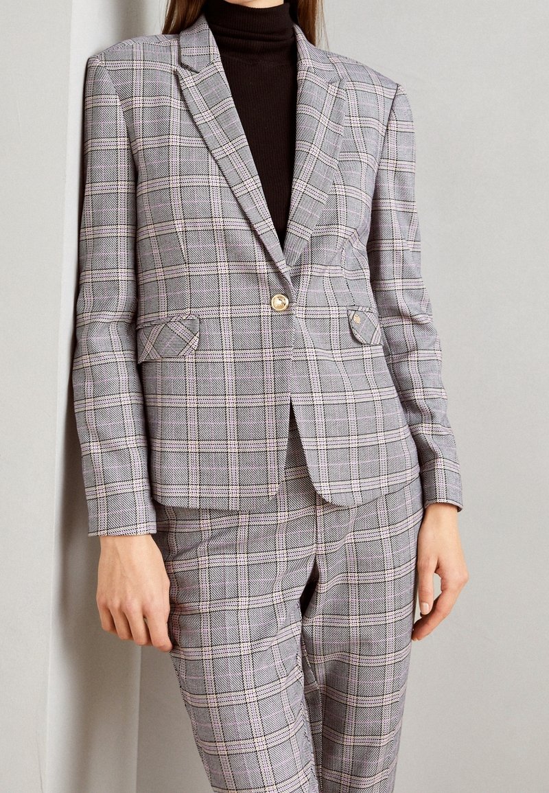 Szara kratkowana marynarka z złotym guzikiem, z kołnierzem z wycięciem i przednimi kieszeniami. Noszona na ciemnym golfie, w zestawie z pasującymi do niej kratkowanymi spodniami.