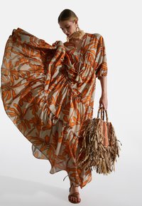 Wellenförmiges Maxikleid aus leichtem Material mit orangen Blumenmustern, gerafften Details und einer Quastenbindung. Kombiniert mit einer fransigen Strohtasche.