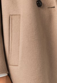 Béžový kabát z vlněné směsi s jemnou texturou, vybavený jedním černým knoflíkem a skrytým bočním kapsou zdůrazňující šitý design.