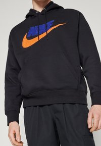 Sudadera negra con bolsillo canguro en la parte frontal. Presenta un logo de Nike en azul brillante y naranja. Está hecha de un tejido texturizado y cuenta con capucha con cordones.