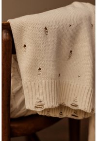 Pull en maille couleur crème avec des trous délibérément usés, drapé sur le dossier d'une chaise en bois sombre.