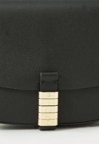 Chiusura di una borsa in pelle nera con cinque barre orizzontali dorate incise con le lettere che formano "PINKO" su una superficie bianca.